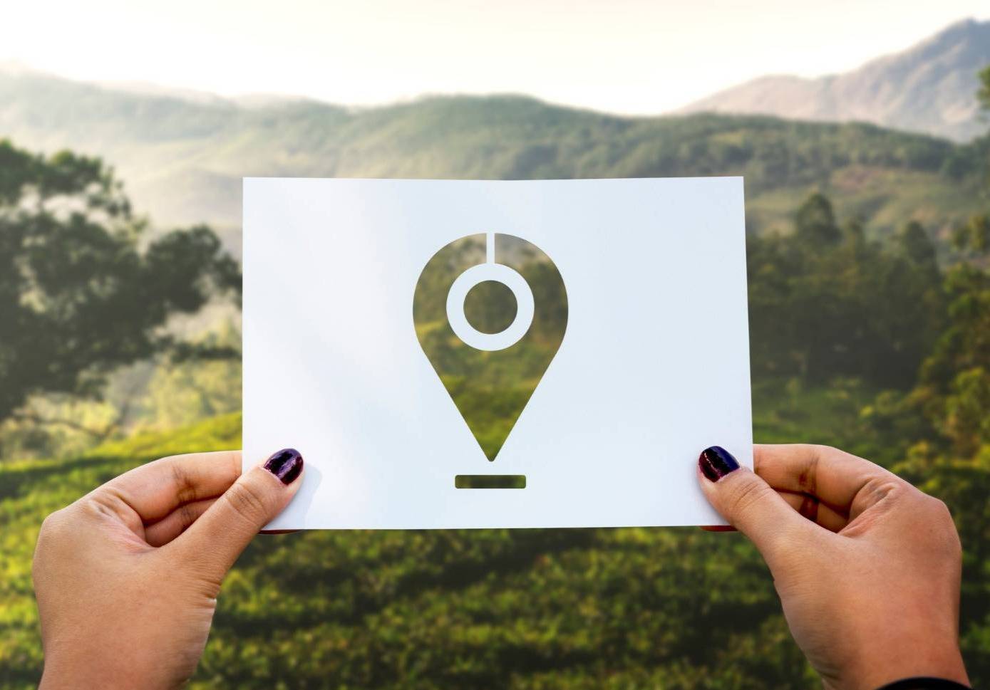 Mãos a segurar uma folha branca com ícone de  localização recortado, com paisagem verde ao fundo