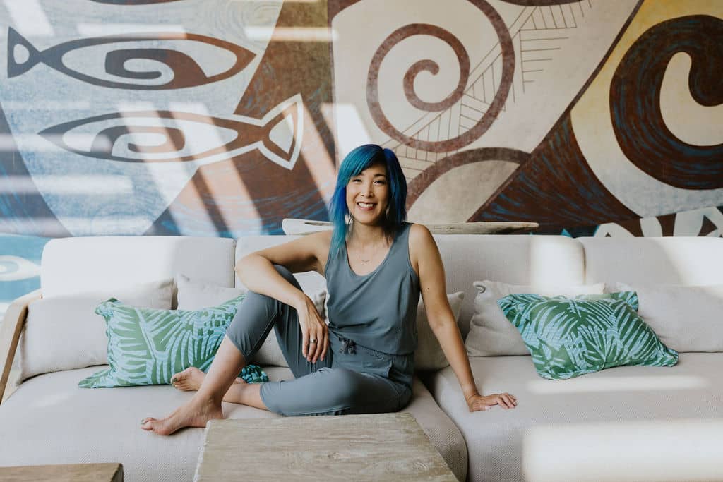 An East Asian woman with blue hair wearing a gray-blue sleeveless jumpsuit sits with her legs and feet on an off-white couch. There are green and blue pillows next to her and there is a mural in the background with stylized fish and swirls in blue, brown and cream.