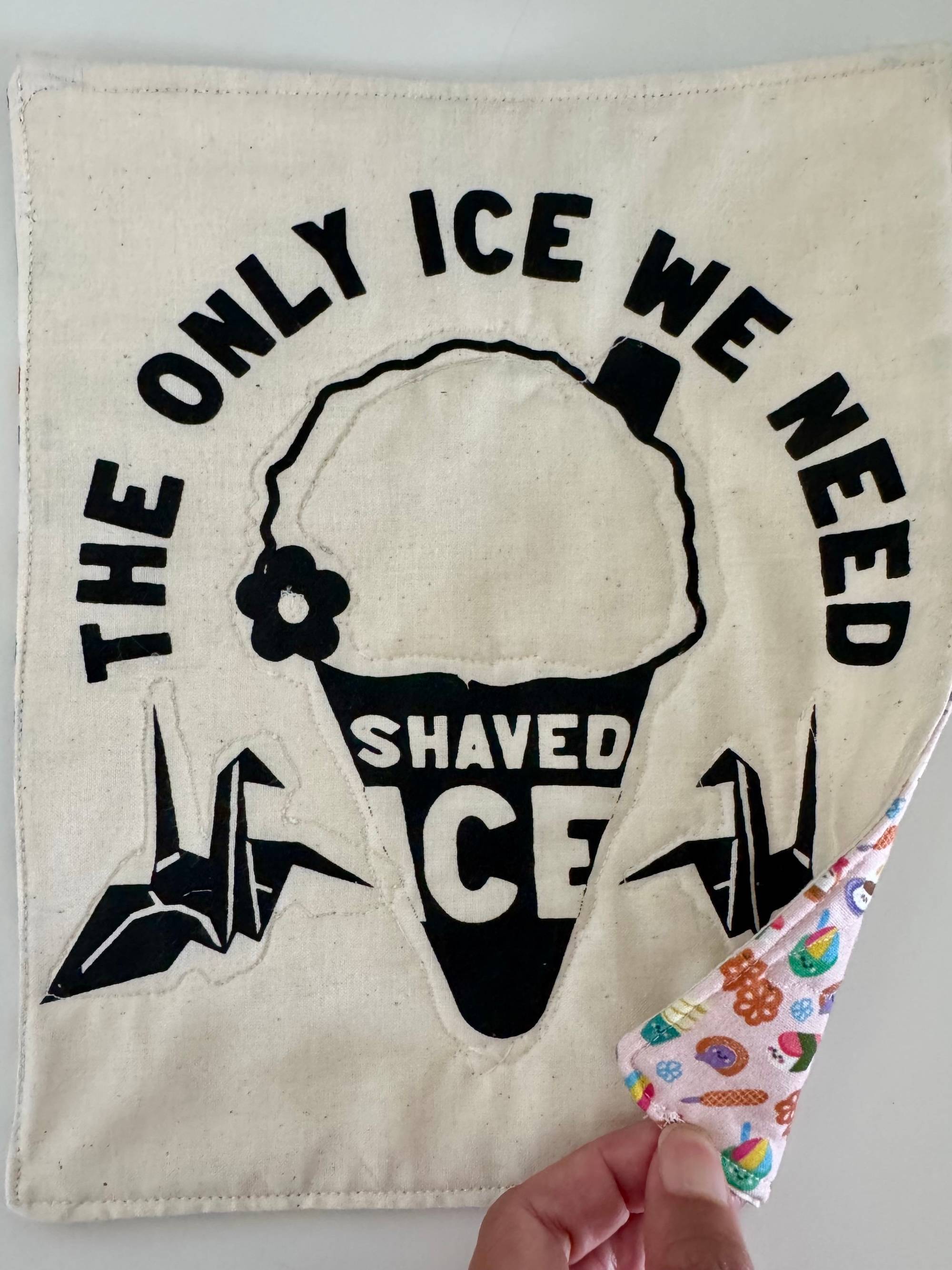 A quilted patch that says the Only Ice we need is shaved ice with a drawing of two origami cranes and a shaved ice cone with a straw and a flower and on the backside of the quilt there's a corner showing of the cute Hawaiian snack fabric pattern.