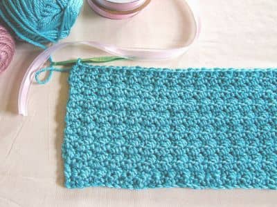 Turquoise crochet swatch with tight, textured stitches displayed on a flat surface. A pink crochet hook and yarn skeins in soft pastel colors are visible at the top left corner.