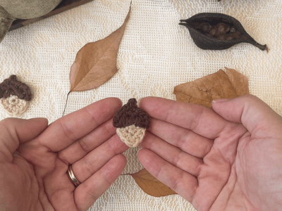 Hands holding a small crocheted acorn appliqué with autumn leaves and yarn in the background.