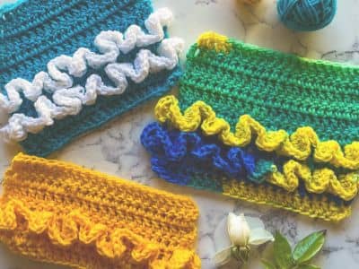 Three colorful crochet swatches decorated with tutu-style ruffles in contrasting yarn colors. A teal swatch features white ruffles, a green swatch has yellow and blue ruffles, and a bright yellow swatch has matching yellow ruffles. Two yarn balls and a wh