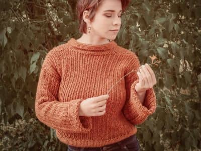 A woman in a textured cinnamon-colored knit sweater stands in front of muted green foliage, gently holding a delicate orange flower and looking down at it, styled in a soft modern vintage French chic aesthetic.