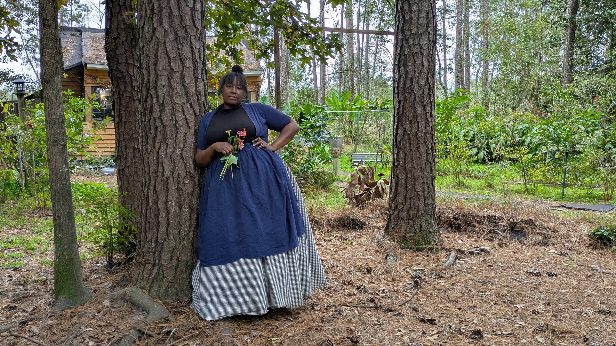 woman in a blue apron and checkered skirt leaning on a tree