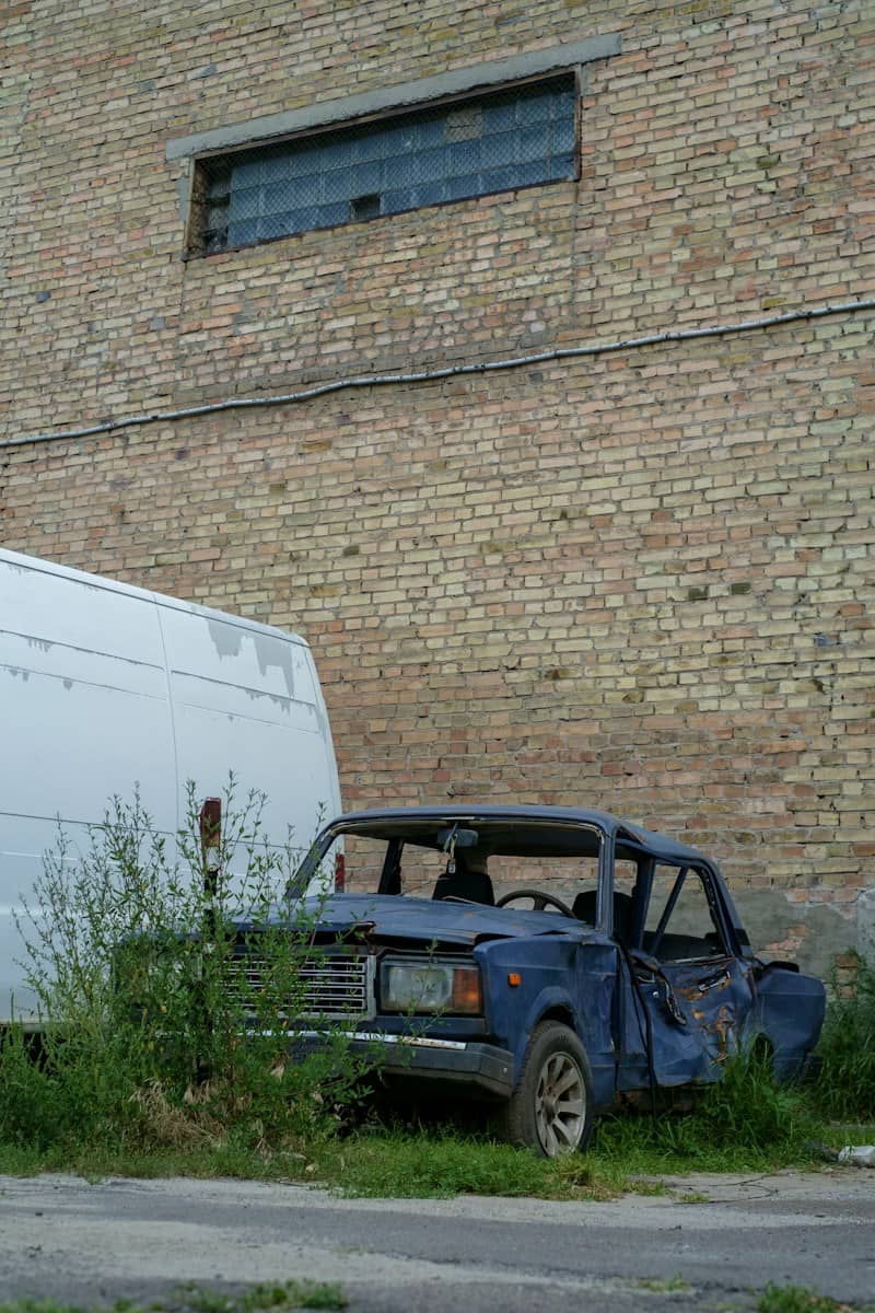 A damaged blue car beside a white van against a weathered brick wall