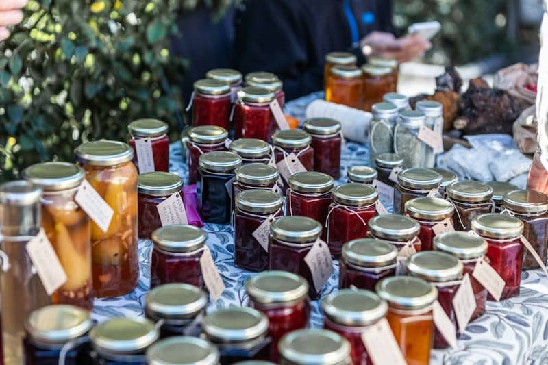 Many glass jars of colorful fruit preserves and jams on a patterned tablecloth