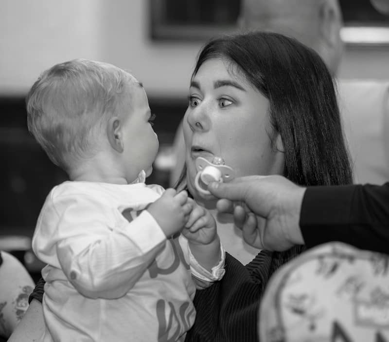 A woman holding a baby who is reaching for a pacifier in her mouth