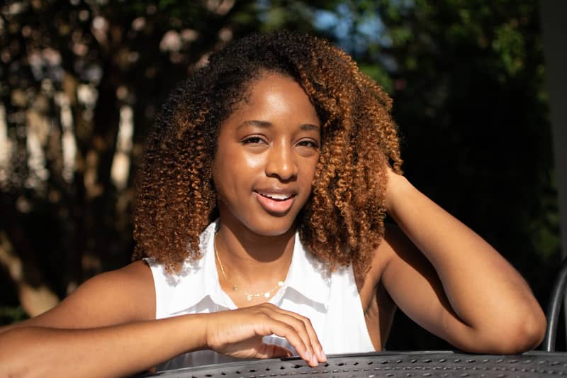 Image of Asia Ohenhen, a woman with reddish brown curly hair, in a white dress. The sun illuminates her face as she rests her ams on a cast iron garden table.