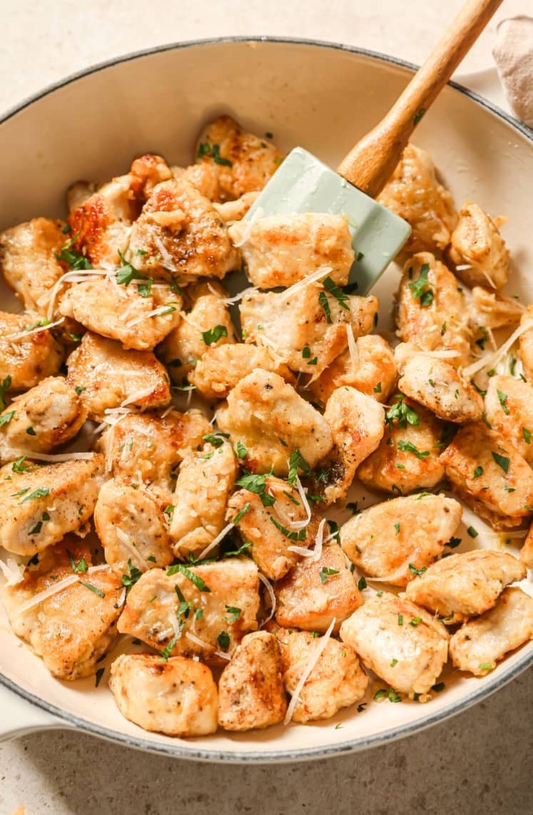 Golden-brown sautéed chicken pieces topped with shredded cheese and chopped herbs in a white skillet, being stirred with a light green spatula.