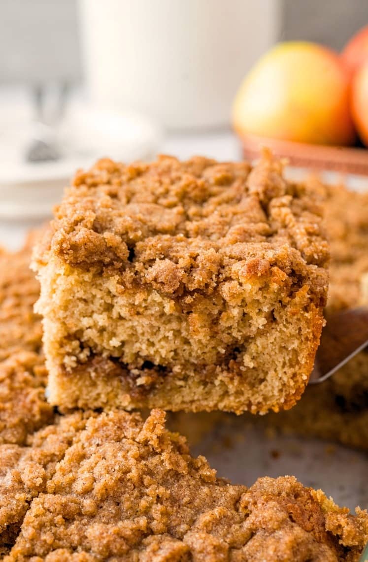 A close-up of a slice of cinnamon coffee cake with a crumbly streusel topping being lifted from a tray.