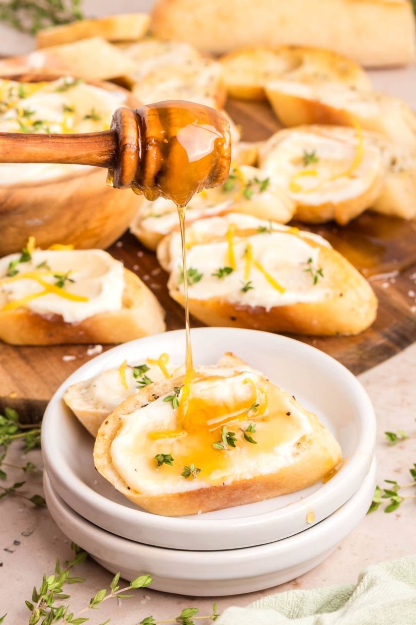 A honey dipper drizzles honey over toasted bread topped with creamy cheese, fresh thyme, and lemon zest. More prepared toasts are visible in the background on a wooden board.