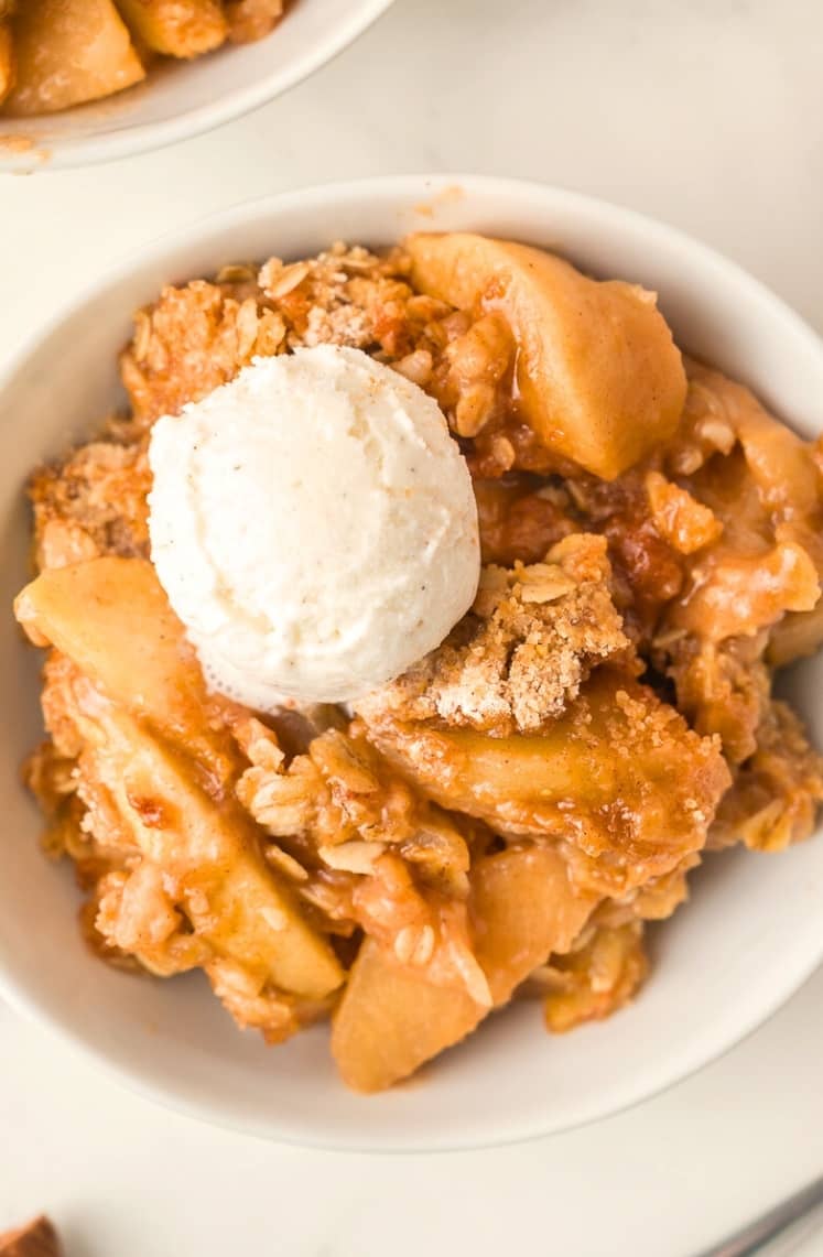 A bowl of apple crisp topped with a scoop of vanilla ice cream, featuring baked apple slices and crumbly oat topping.