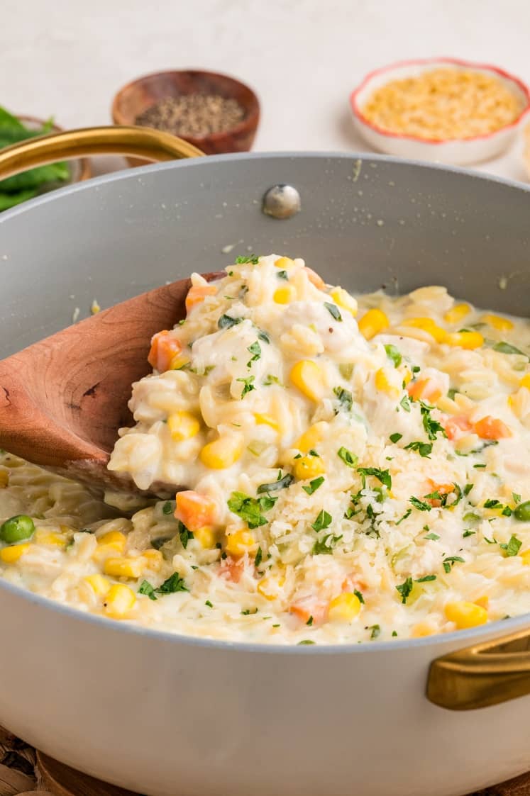 A creamy orzo pasta with corn, carrots, green peas, and herbs is lifted from a pan with a wooden spoon. Parmesan cheese and seasonings are visible in the background.
