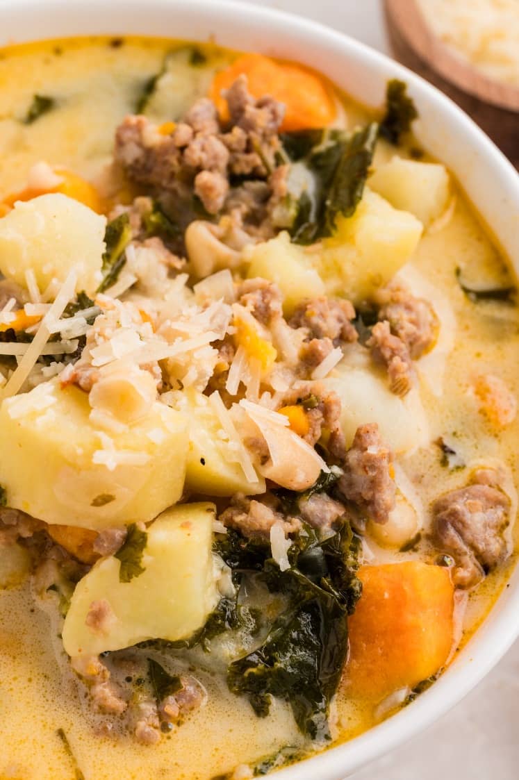 A close-up of a bowl of soup containing ground meat, potatoes, kale, white beans, carrots, and topped with grated cheese.