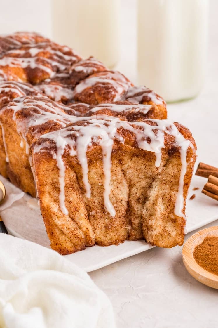 A loaf of cinnamon pull-apart bread with icing drizzled on top, with one piece partially pulled away; two bottles of milk are in the background.