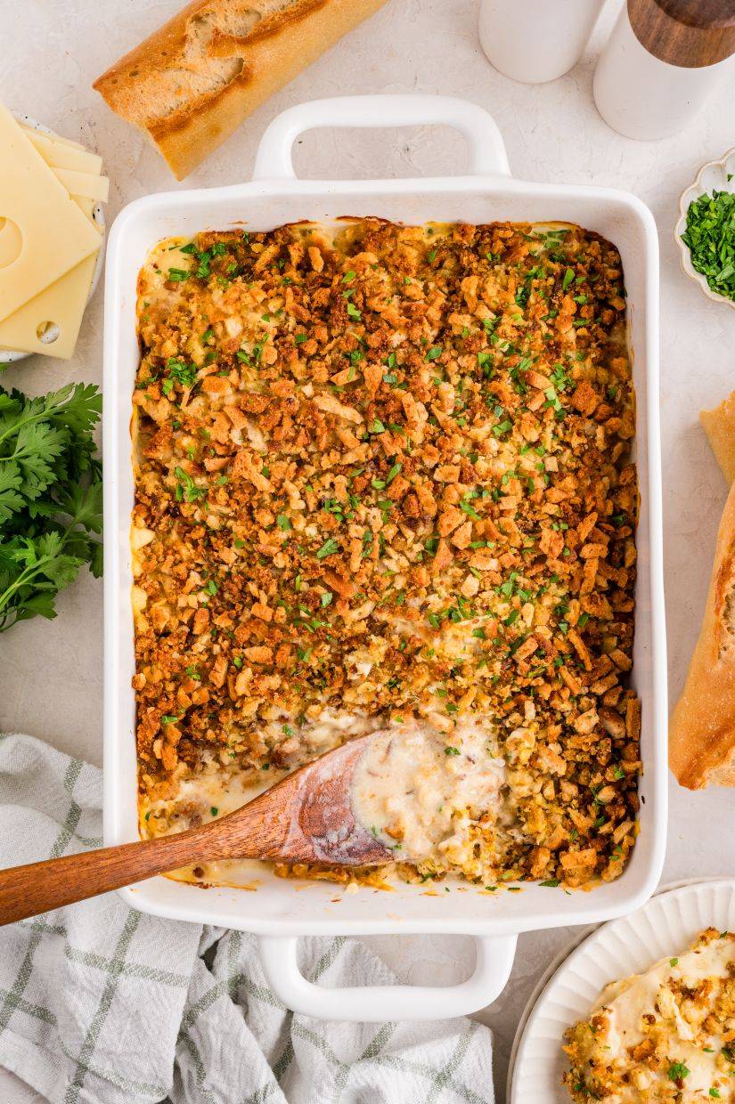 A baked casserole topped with golden, crispy crumbs and chopped herbs in a white dish, with a wooden spoon scooping out a creamy serving. Slices of bread, cheese, and fresh parsley are nearby on the table.