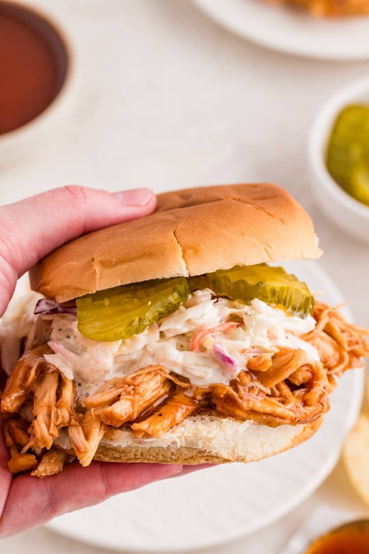 A hand holding a pulled chicken sandwich topped with coleslaw and pickle slices on a hamburger bun.