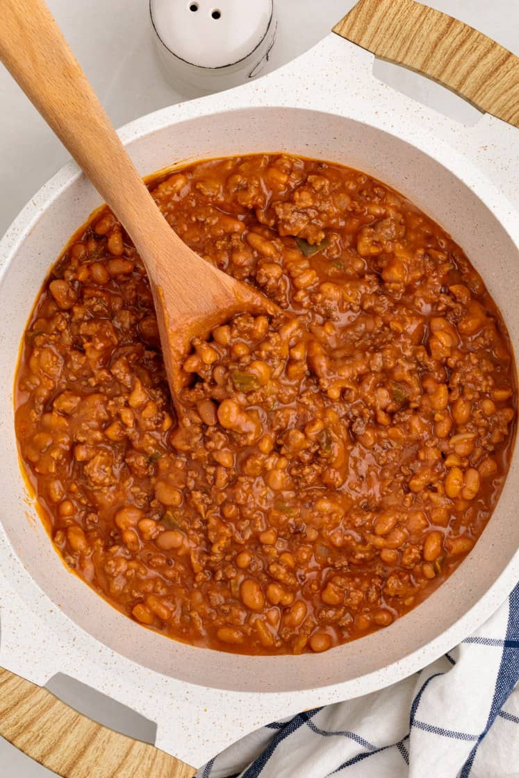 A white Dutch oven filled with baked beans and ground beef, with a wooden spoon resting inside and visible specks of seasoning in the red sauce.