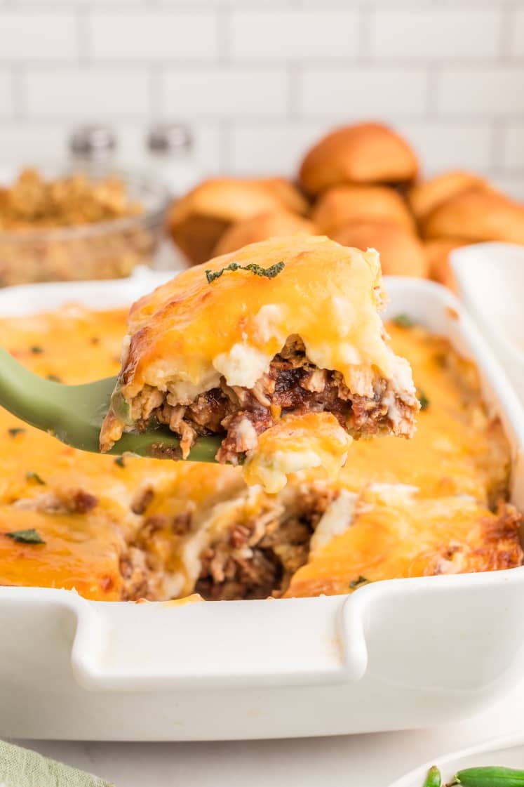 A close-up of a wooden spatula holding a slice of cheesy baked casserole with shredded meat, beans, and sauce, above a white baking dish.