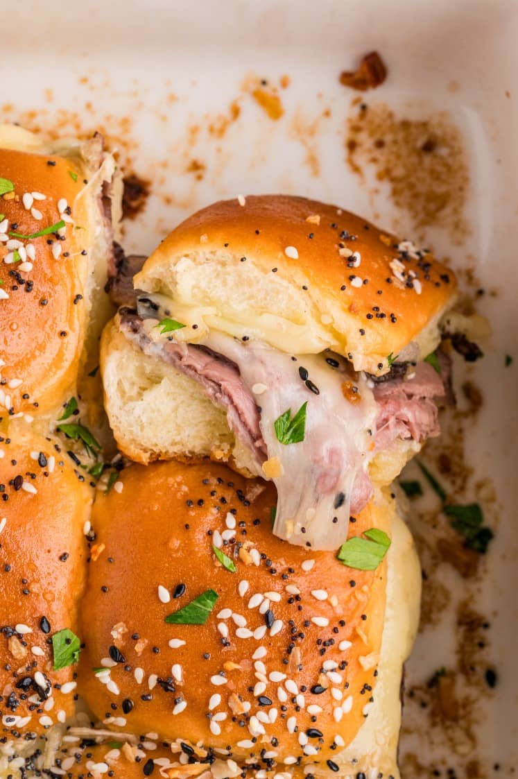 Close-up of baked sliders with golden buns, melted cheese, roast beef, and sprinkled sesame and poppy seeds, garnished with fresh herbs.