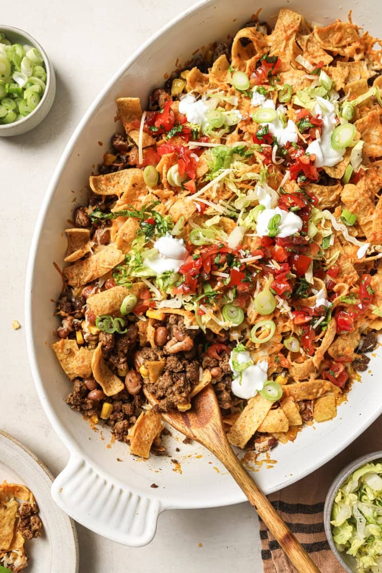 A white baking dish filled with taco casserole topped with Fritos, ground beef, corn, beans, shredded cheese, lettuce, diced tomatoes, green onions, and dollops of sour cream. A wooden spoon rests in the dish.