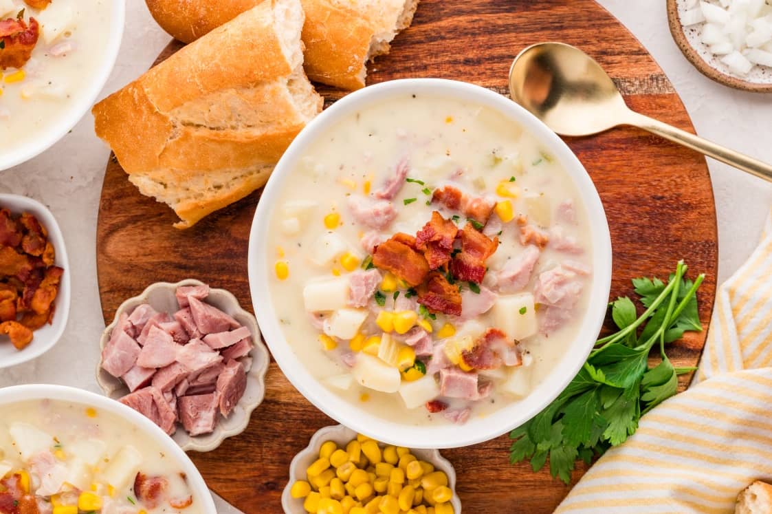A bowl of creamy soup with ham, potatoes, corn, and bacon bits sits on a wooden board, surrounded by French bread, parsley, chopped ham, and corn, with a gold spoon nearby.