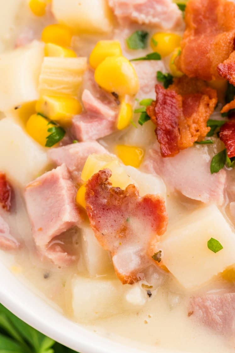 A close-up of a spoonful of creamy soup with diced potatoes, corn, ham, and a piece of crispy bacon, held above a bowl filled with the same soup.