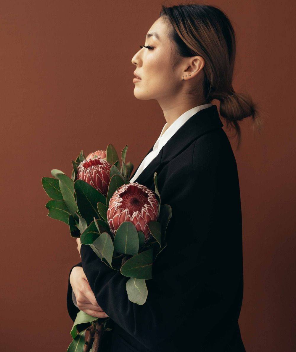 In a world of hustle and bustle, this empowering image captures a moment of calm confidence. An asian woman in a stylish suit, holding flowers, exudes elegance and grace against a warm brown backdrop. Her closed eyes and thoughtful gaze hint at inner wisdom and determination. #elegance #strength #serenity #confidence #empowerment