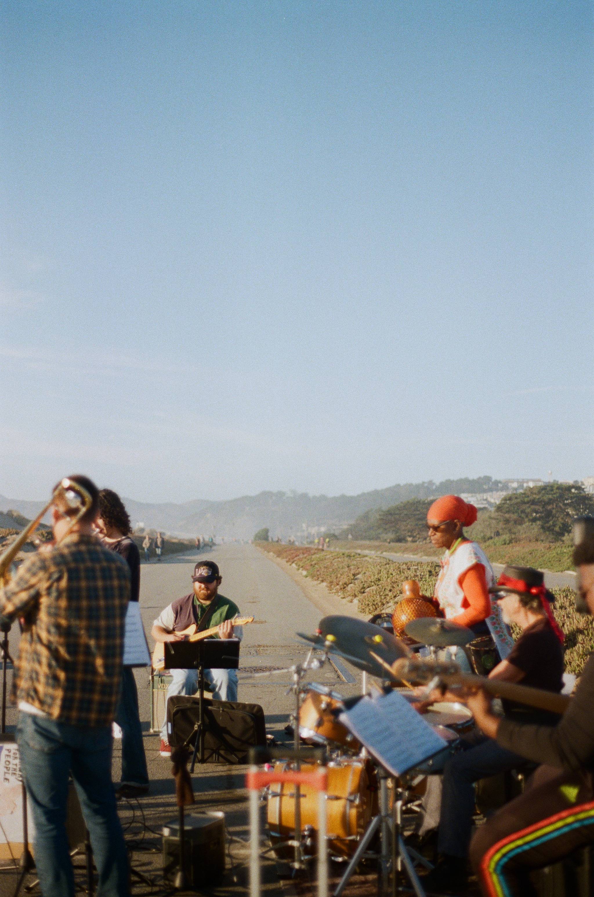 A group of musicians performing outdoors on a path surrounded by greenery and hills in the background. The ensemble includes a guitarist, a drummer, and other instrumentalists. One musician is playing a drum set, while another stands holding a percussion instrument. The atmosphere is relaxed and sunny under a clear blue sky.