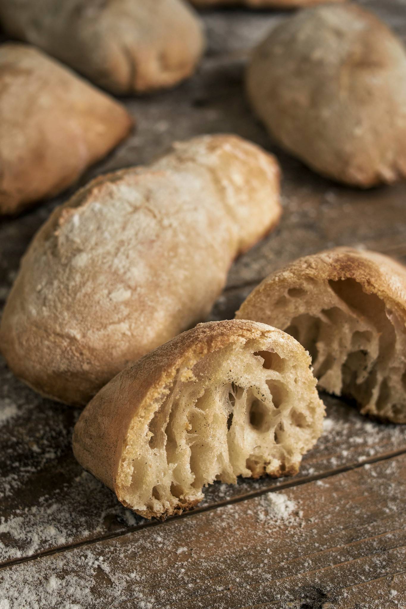 Rustic no-knead ciabatta rolls on a floured wooden surface, with one roll cut open to show the airy crumb.