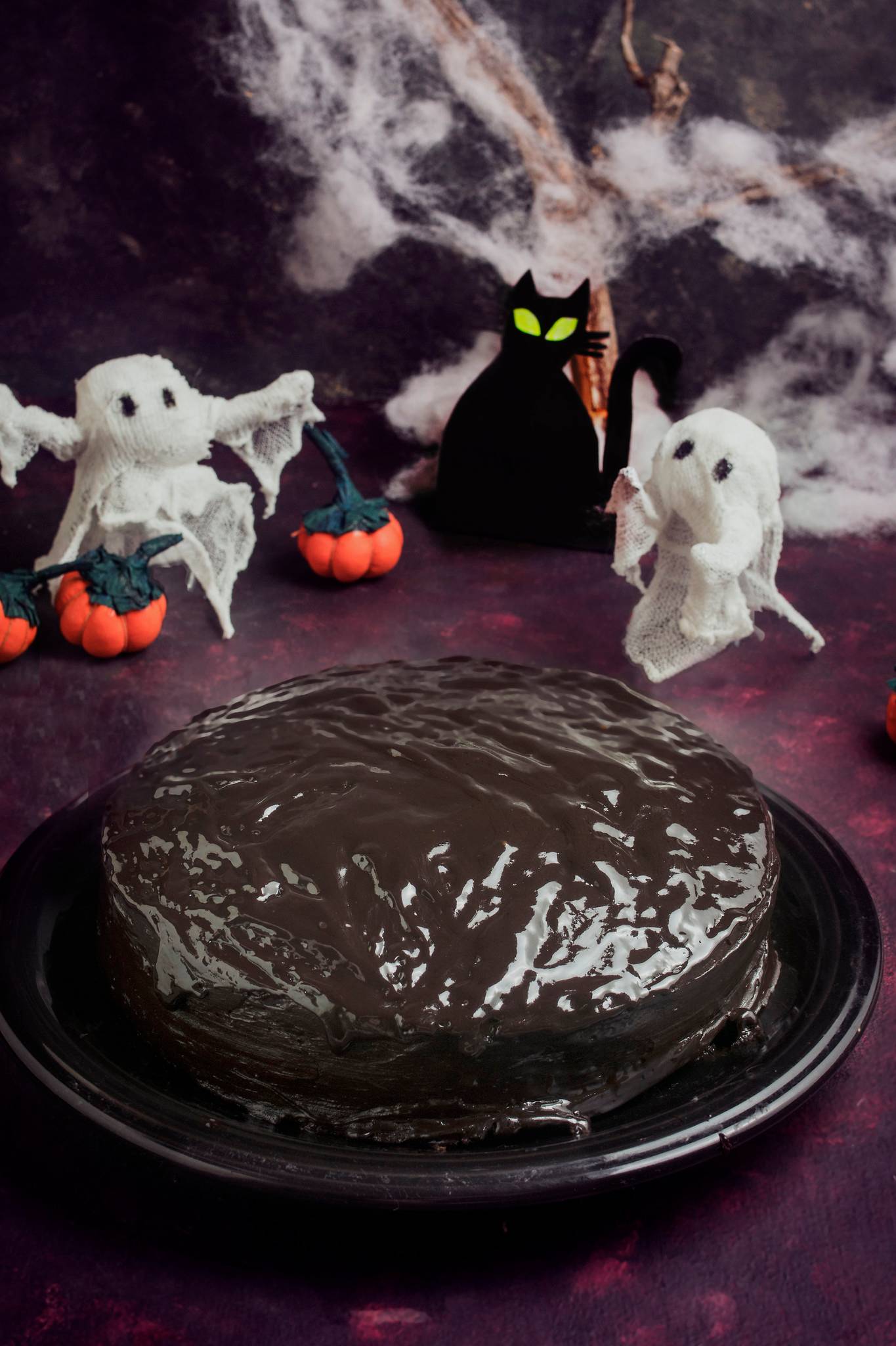 Rich chocolate Halloween cake with glossy chocolate icing, surrounded by ghost decorations, miniature pumpkins, cobwebs and a black cat ornament.