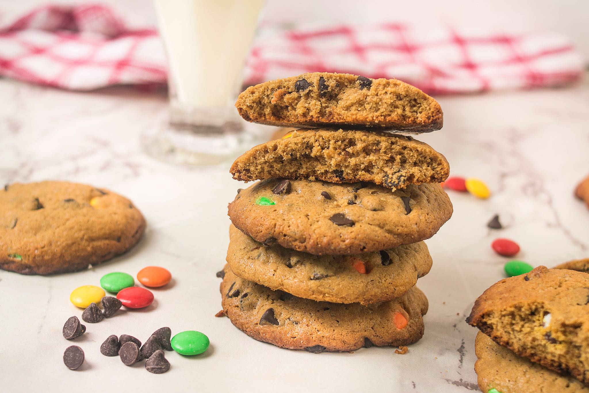 tack of browned butter chocolate chip cookies, with broken cookies showing the soft centre, surrounded by chocolate chips and colourful candy-coated chocolates.