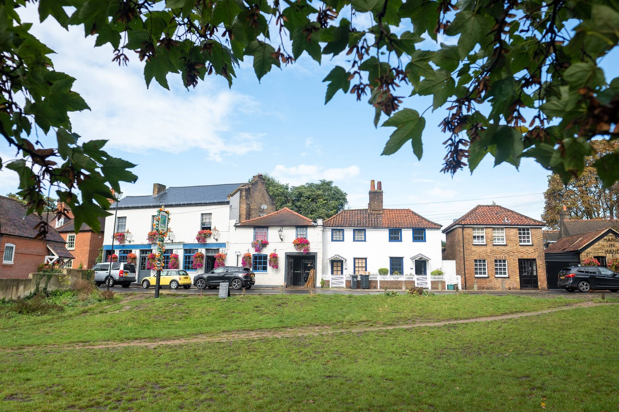 The Hand in Hand and Crooked Billet pubs beside Wimbledon Common, with whitewashed buildings, red-tiled roofs and leafy branches framing the village green.