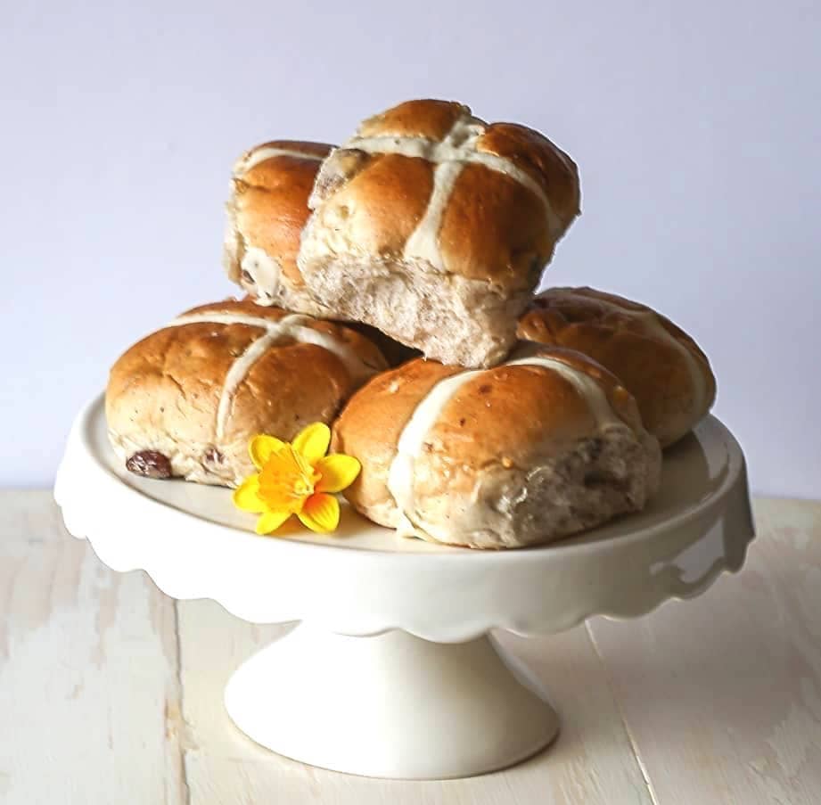 Traditional hot cross buns stacked on a white cake stand with a small yellow spring flower