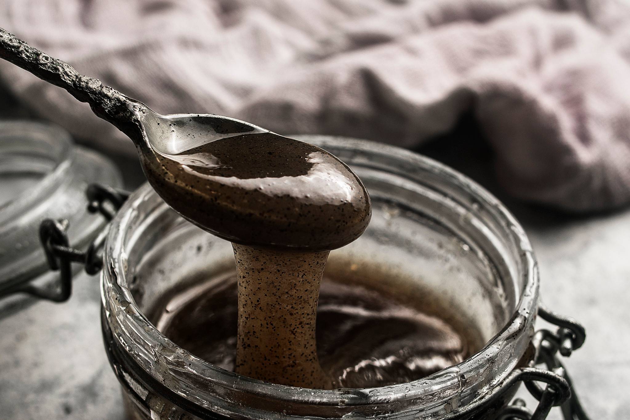 Thick homemade vanilla bean paste dripping from a spoon into a glass jar, with visible vanilla seeds.