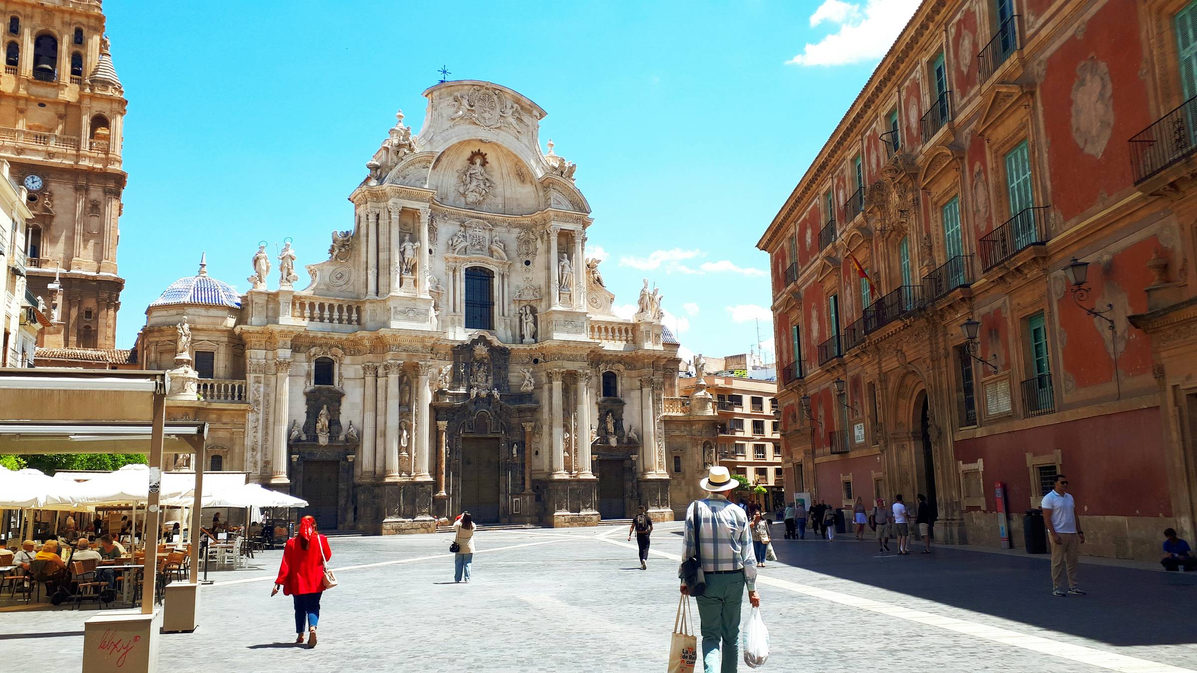 Murcia Cathedral and the surrounding plaza on a sunny day, with people walking across the square and historic buildings framing the scene.
