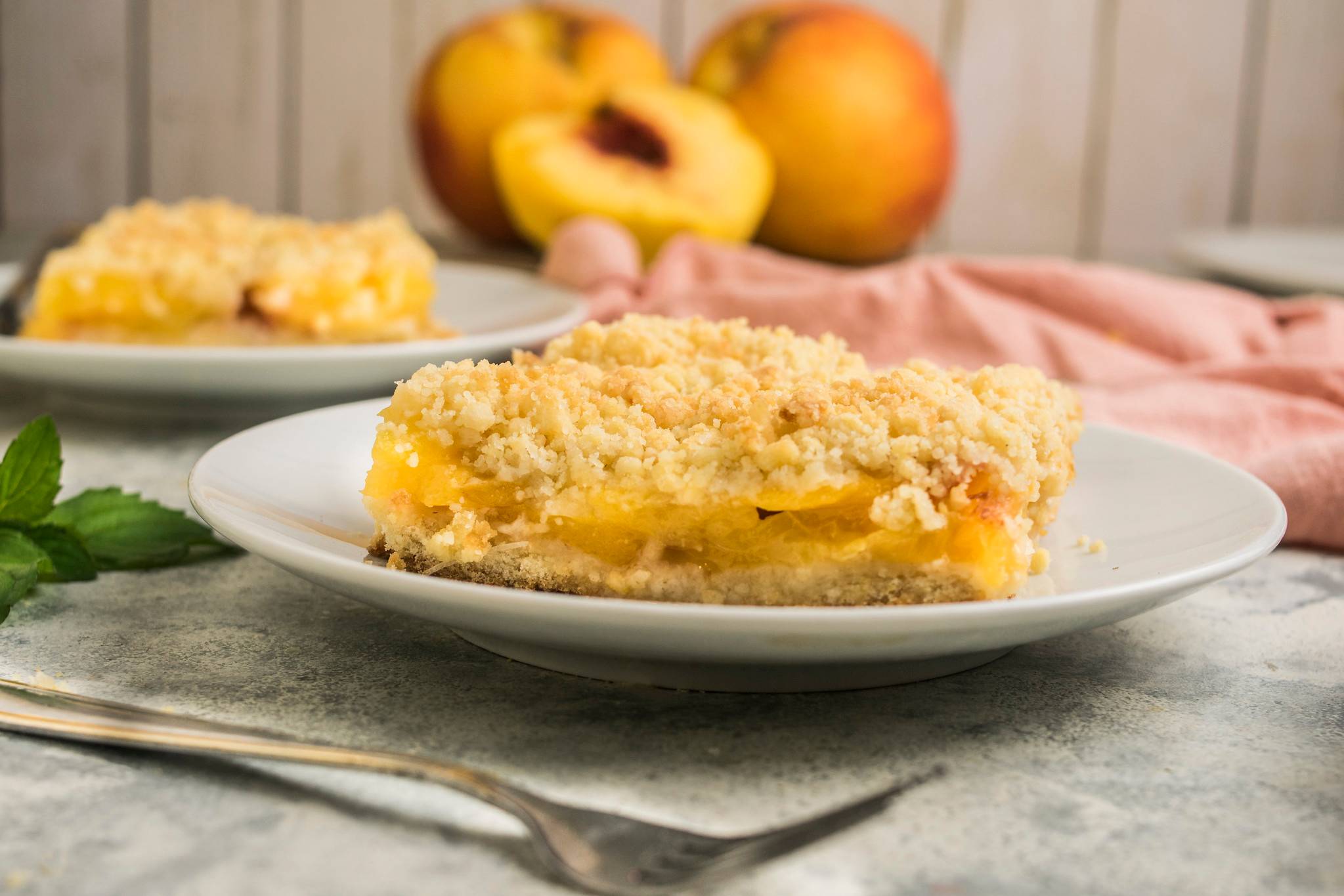 A slice of peach sbriciolata with golden crumble topping and soft peach filling, served on a white plate with fresh peaches softly blurred behind.
