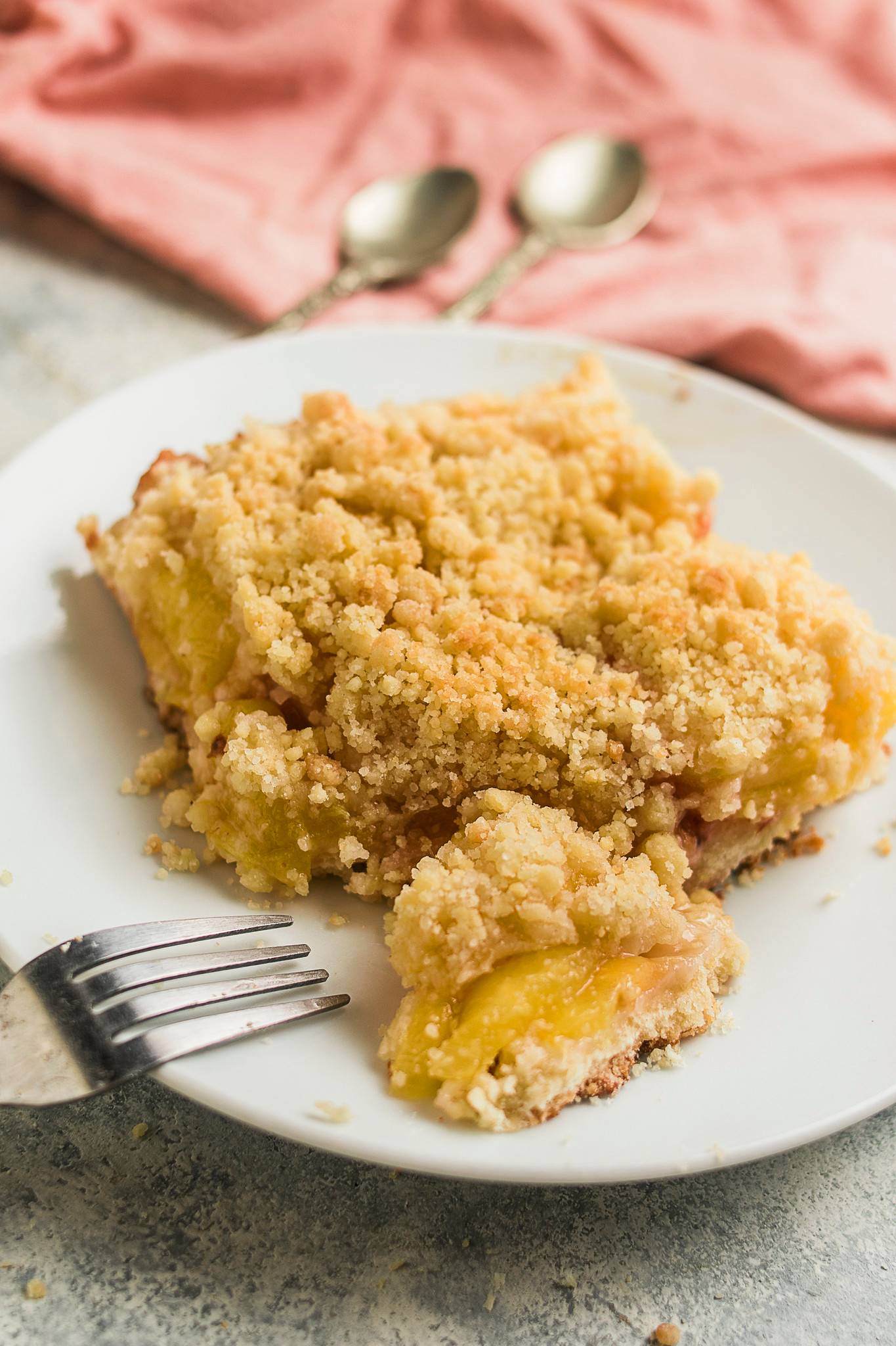 A close-up slice of peach sbriciolata with golden crumble topping and soft peach filling, served on a white plate with a fork ready beside it.