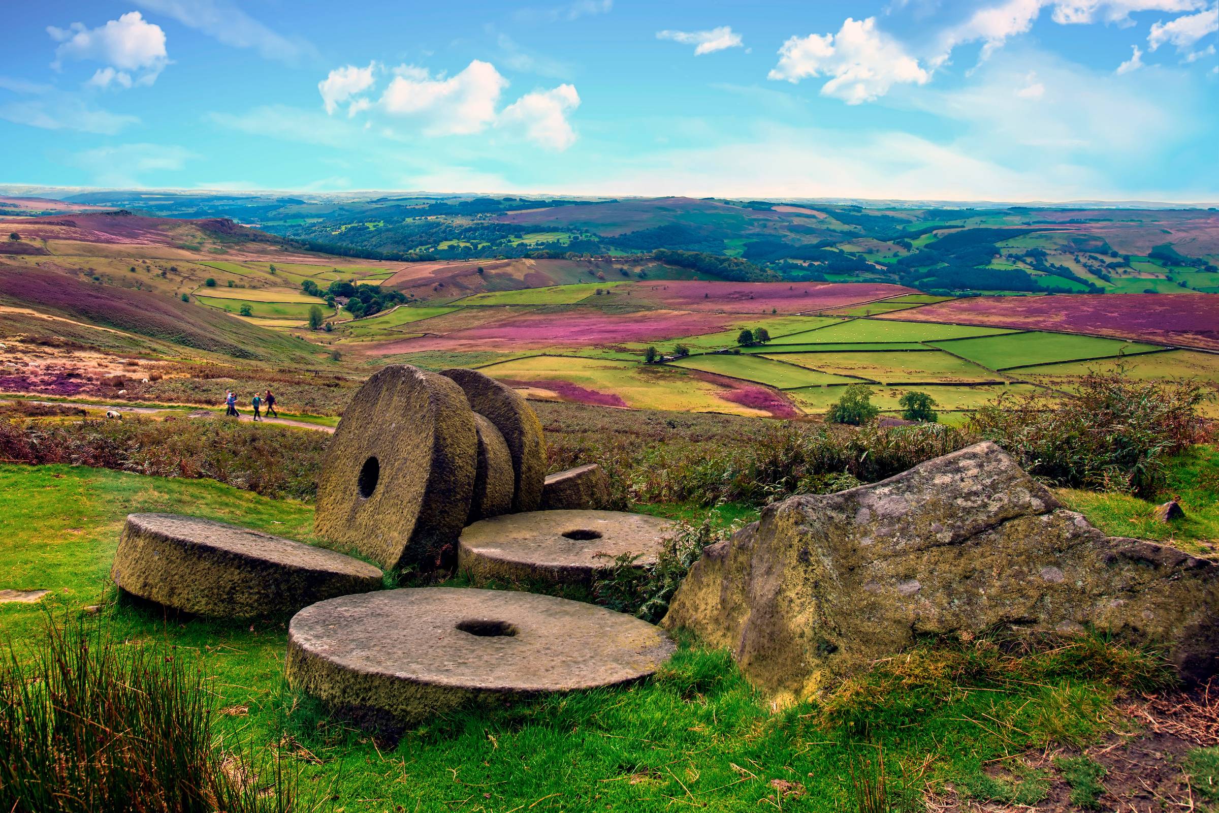 Old millstones on Stanage Edge in the Peak District, with sweeping views across green fields, purple heather and rolling countryside beneath a bright summer sky.