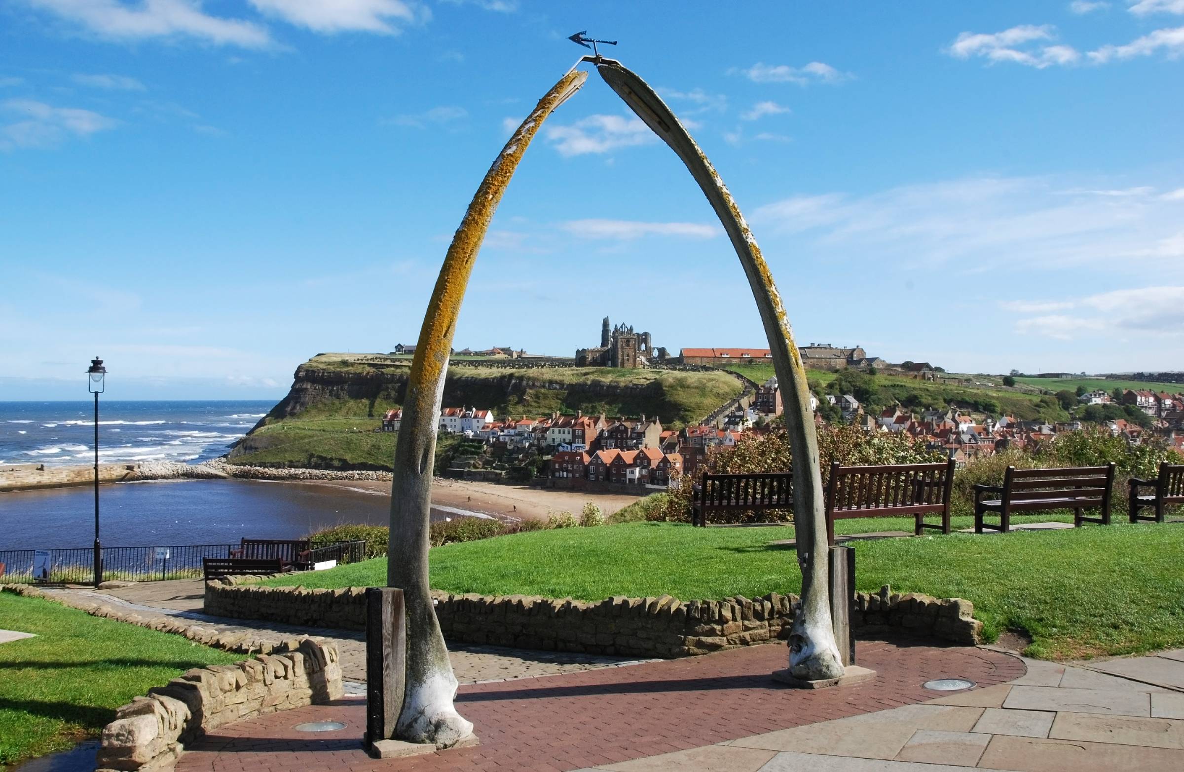 Whitby Abbey framed by the whale bone arch on West Cliff, looking across the harbour, beach and red-roofed houses beneath a clear blue sky.