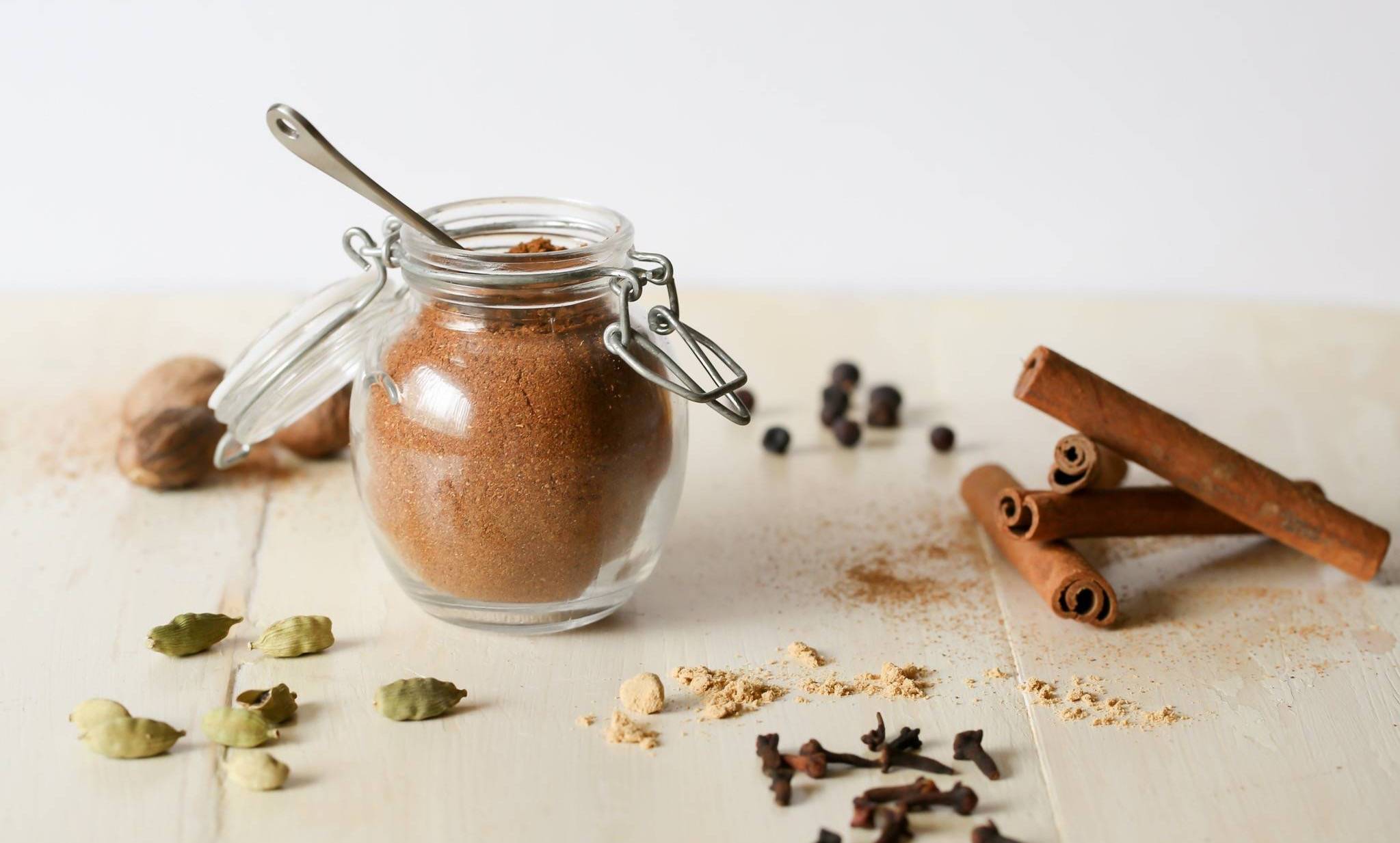 Homemade autumn spice blend in a glass jar, surrounded by cinnamon sticks, cardamom pods, cloves and whole spices on a pale wooden surface.