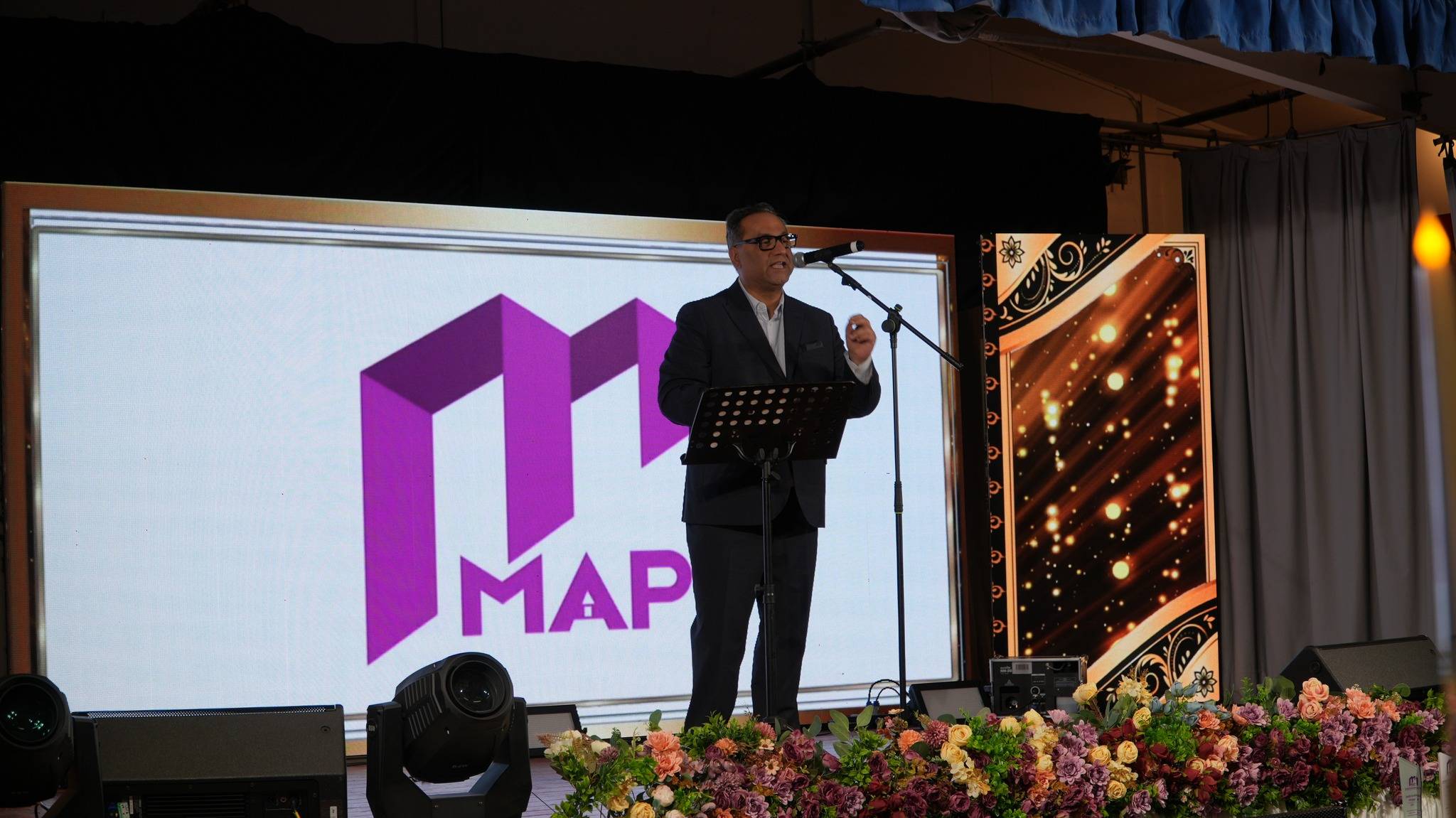A man in a dark suit speaks at a podium on a stage decorated with flowers, positioned in front of a large screen displaying a purple "MAP" logo-The Noble Sunday 