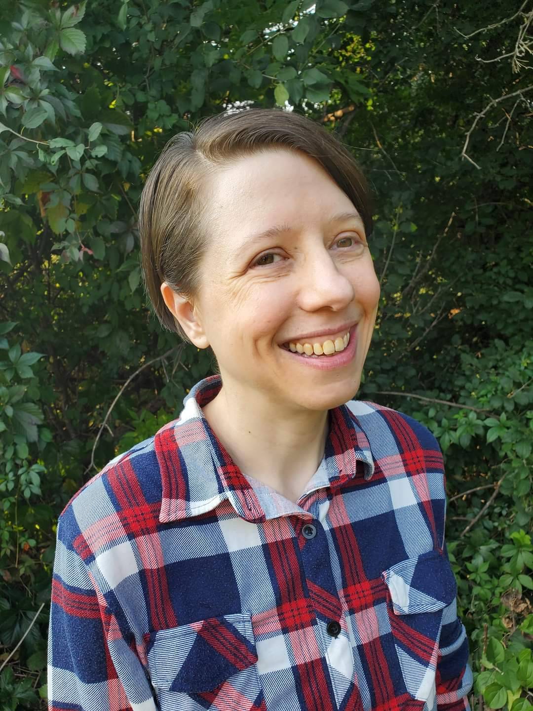 Headshot of Julie Nowak, a smiling white person with short brown hair and plaid shirt in front of greenery outdoors.