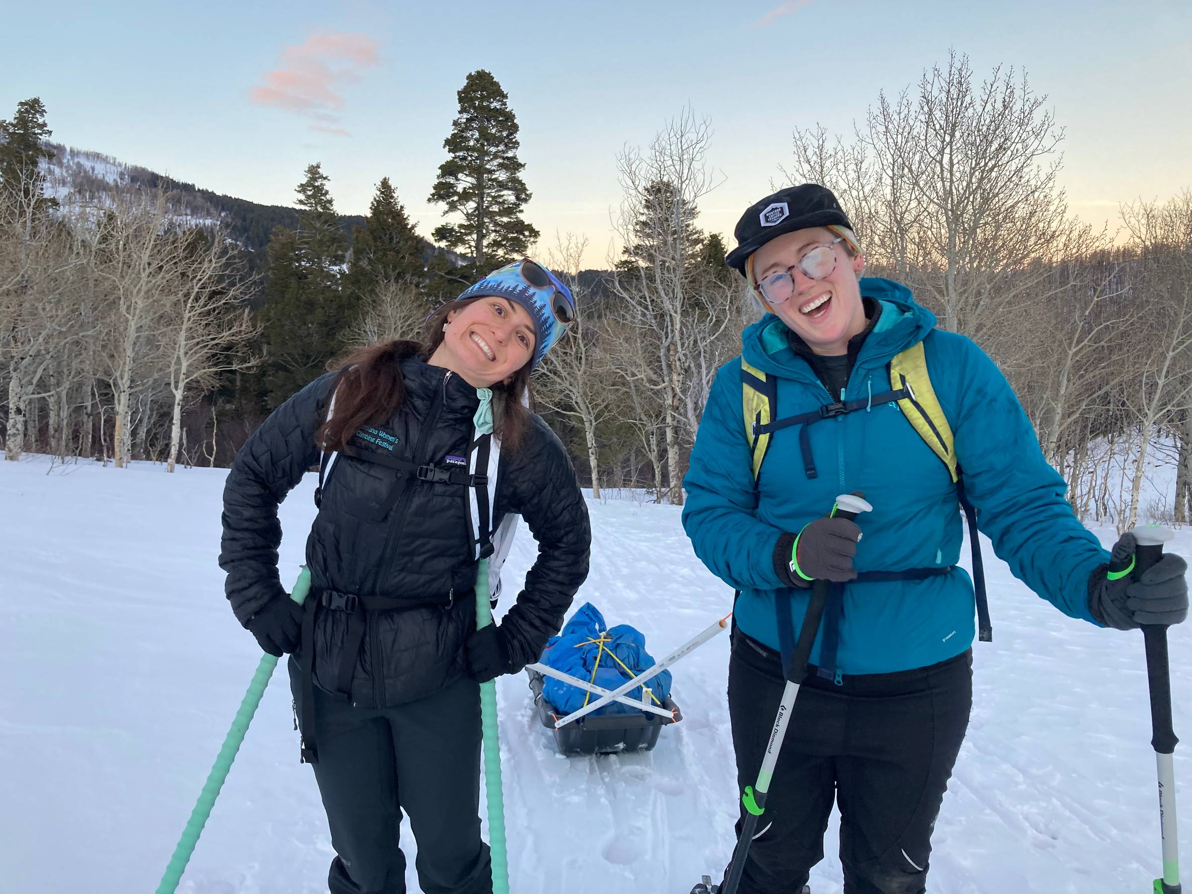 Two girls smiling at the camera on skis and pulling a sled at dusk. Trees in the background.