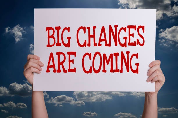 Two hands hold up a white sign on which "Big Changes are Coming" is written in large red letters. The background is a blue sky with white fluffy clouds.
