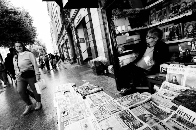City street newsstand with a vendor and passing pedestrians.