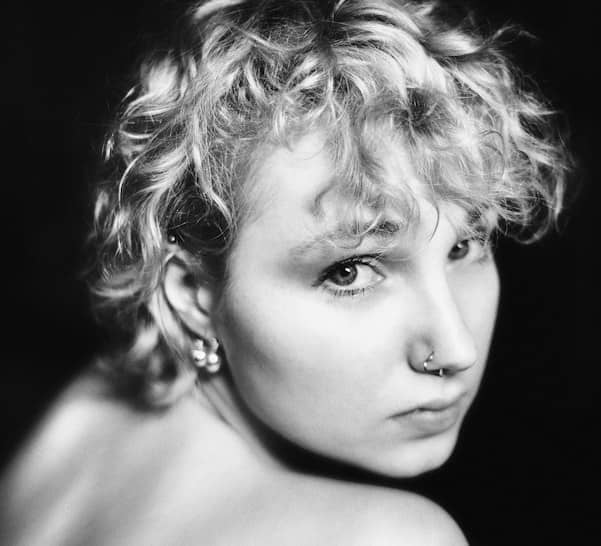 A person with short curly blonde hair and a nose ring looking back