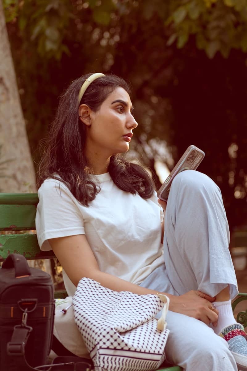 A woman in a white t-shirt holding a phone on a green park bench
