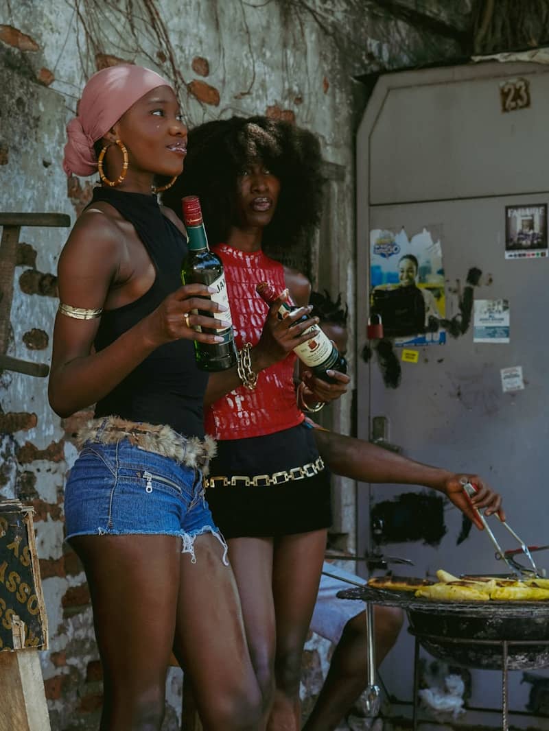 Two women hold bottles while another person grills food.