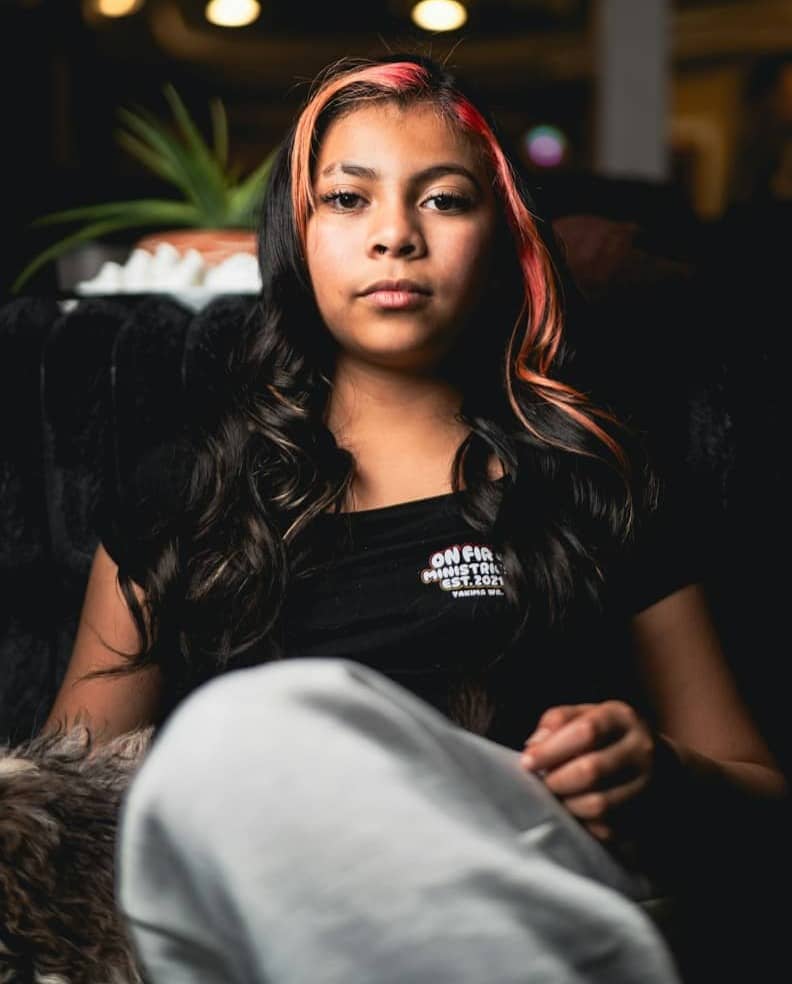 A young person with pink-streaked dark hair in a black t-shirt sitting indoors